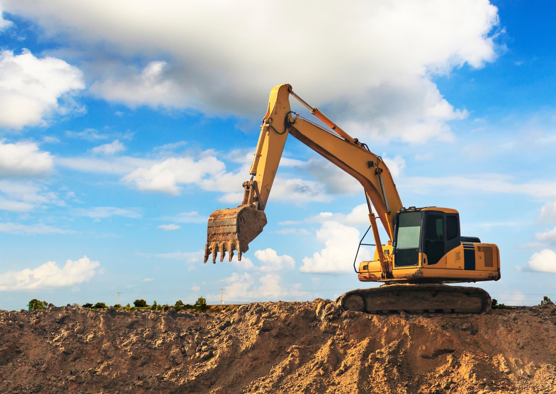 Excavator with a raised bucket standing on a mound of earth against a blue sky